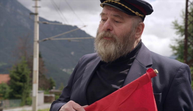 Eine Szene aus dem Film über Werner Pirchner: Wir sehen Erwin Steinhauer im Kostüm eines Bahnwärters. Er steht an einem kleinen Bahnsteig in Tirol, schwenkt eine Rote Fahne und singt.