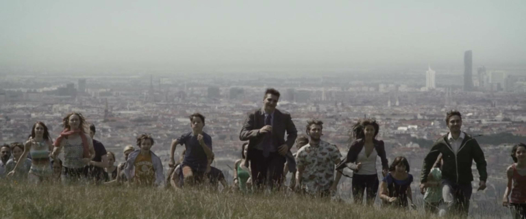 Wir sehen eine Gruppe von Menschen, die gemeinsam einen Hügel hinaufrennen. Hinter ihnen ist eine Großstadt zu sehen.